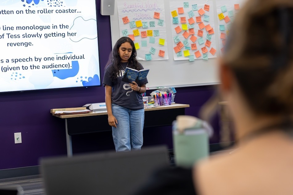 A teen girl reads out loud from a notebook in her hands in front of a classroom of peers 