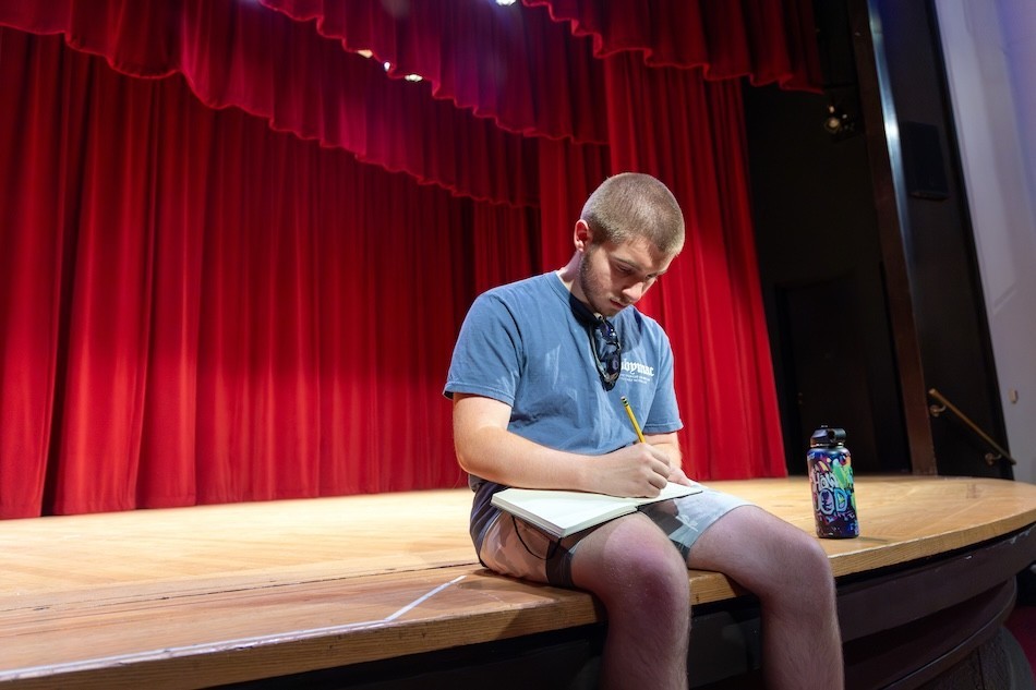 A teen boy sits on the edge of a theatre stage writing in a notebook on his lap
