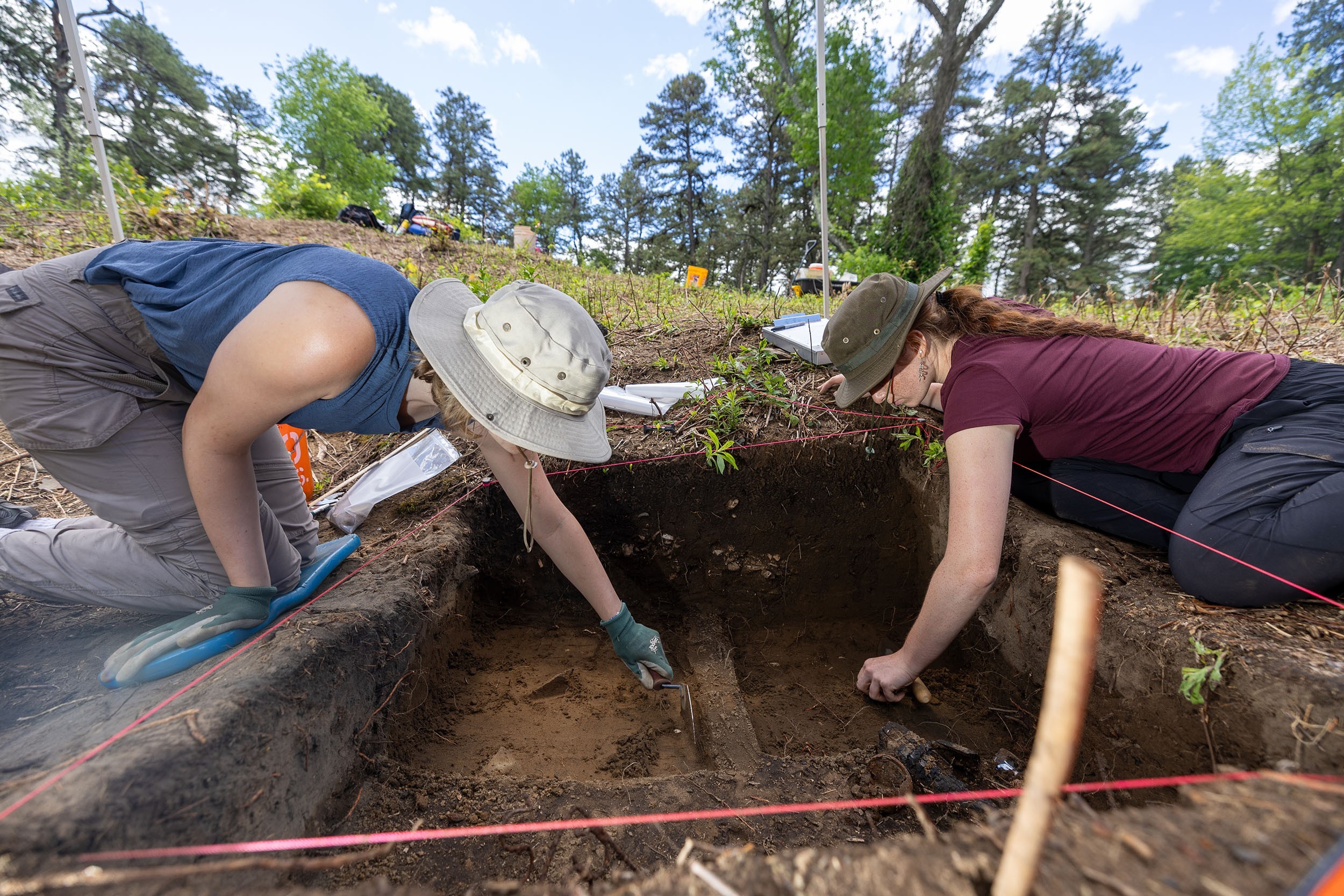 two young women map grid lines in a square of dirt at an archeological field school in Albany's Pine Bush as they reach into the square.