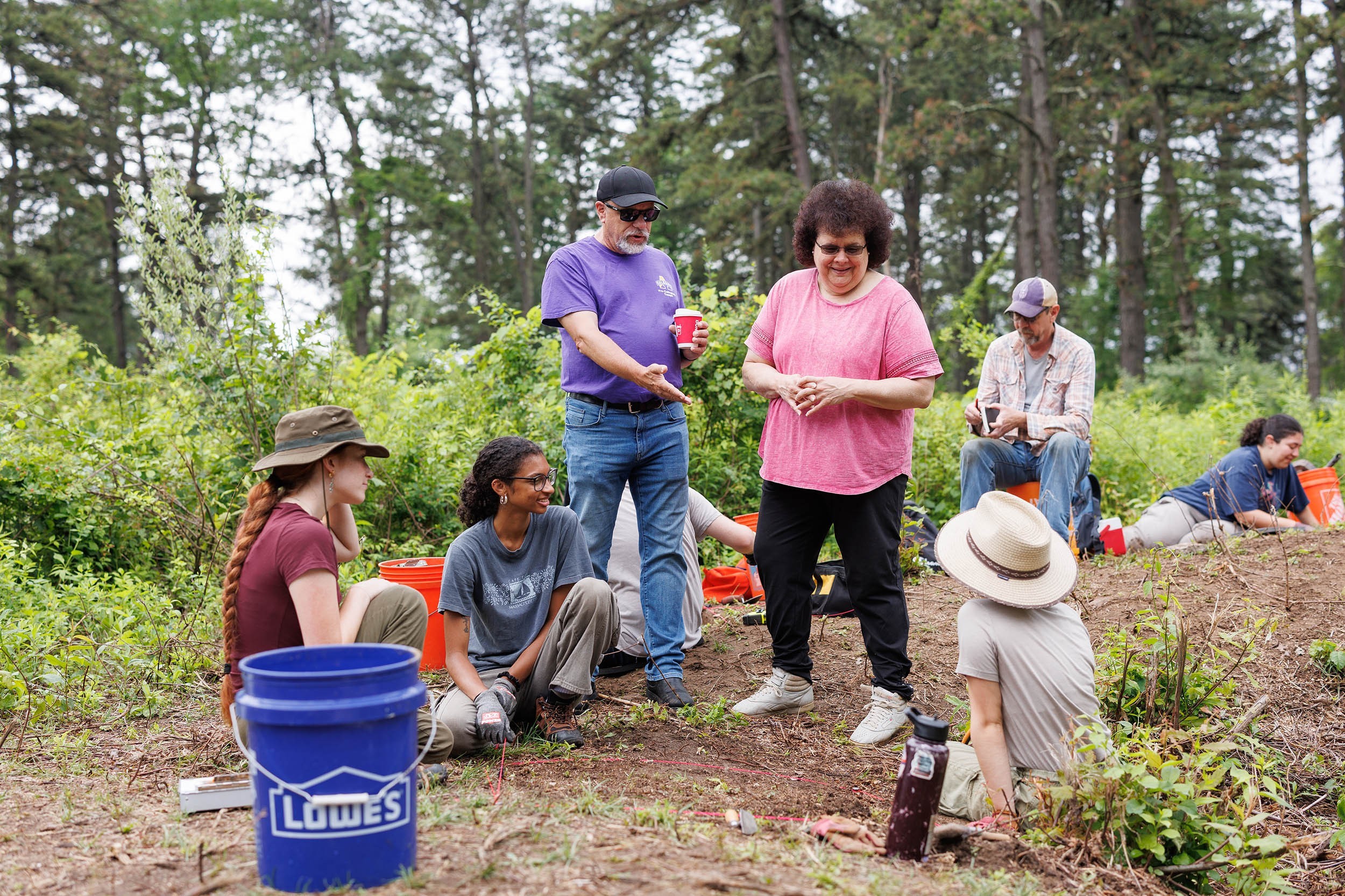 At an archeological field school in the Pine Bush of Albany two students sit on the ground while two faculty members stand and talk to them near buckets and a grid pattern mapping the site.