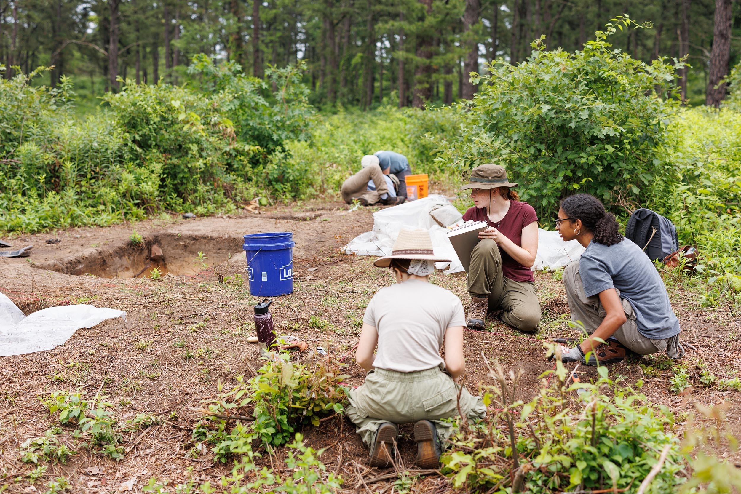 At an archeological field site in Albany's Pine Bush three young women wearing cargo pants kneel over a dirt grid.