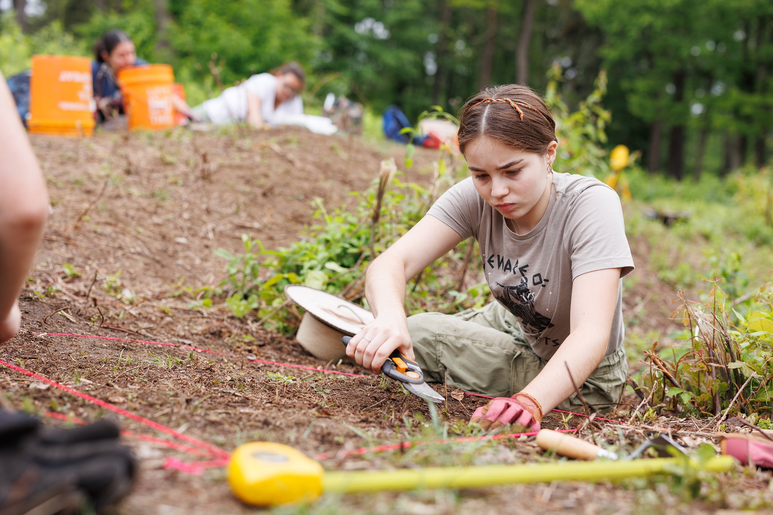 A young woman measures with a measuring tape as she kneels on the ground in khakis at an archeological field school in the woods of Albany's Pine Bush.