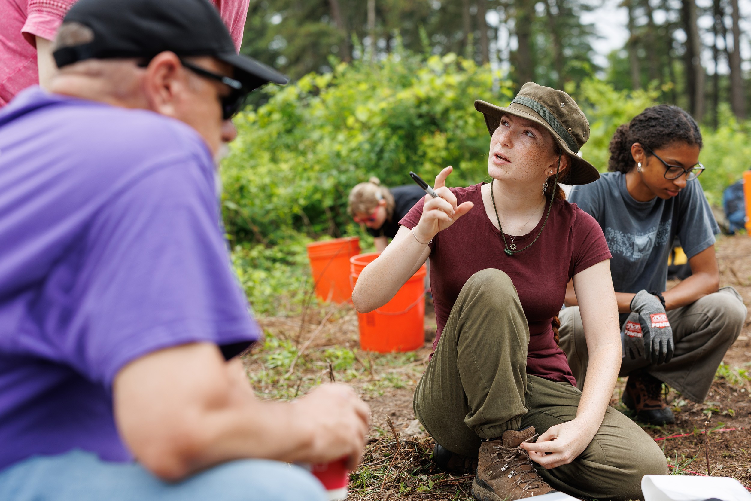 A young college student kneeling in cargo pants and a dark red shirt speaks to two faculty members at Albany's Pine Bush at an archeological field school.