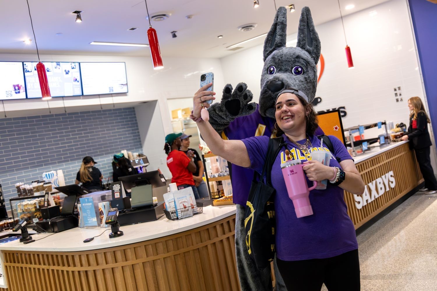 A young woman in a purple shirt stands with a great dane mascot in front of a cafe wearing UAlbany shirts.