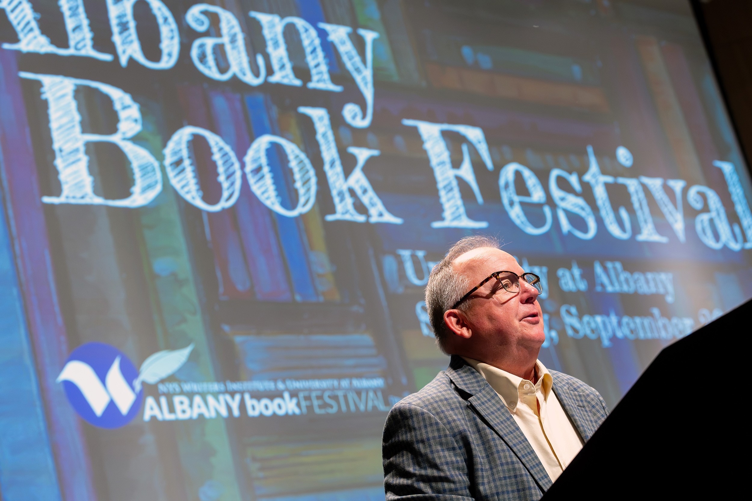 A man speaks at a podium with the words Albany Book Festival behind him on a large screen.