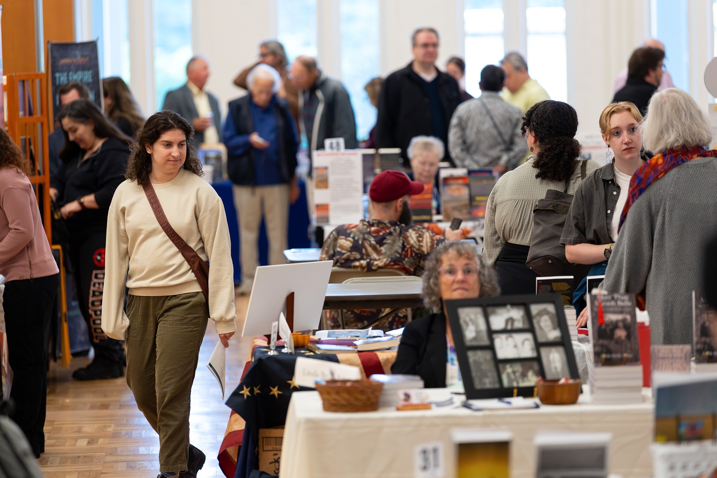 A large group of people move between aisles of people seated with their books during a book show in an auditorium