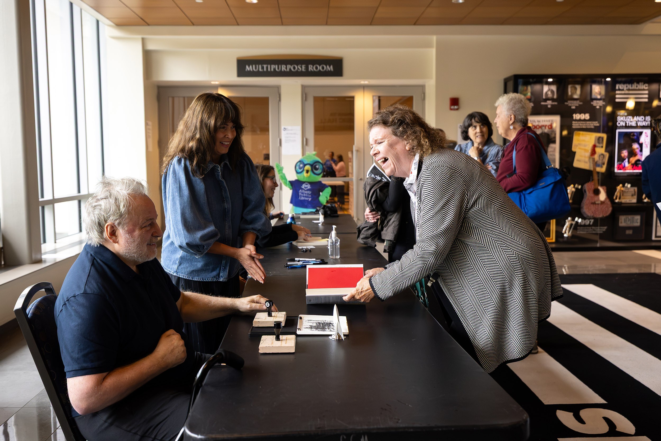 A man is seated signing books as several people line form to get autographs and a woman leans over a book she has placed in front of him.