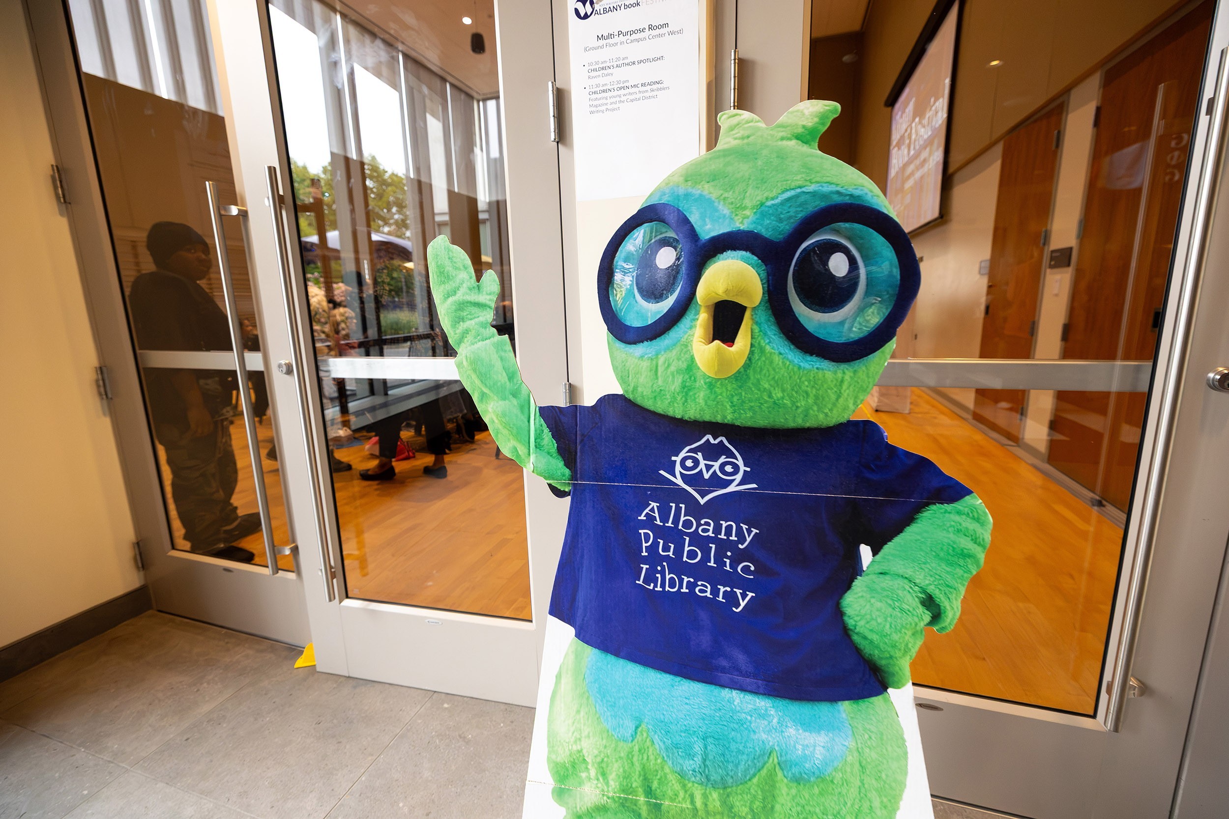 A green owl sign wearing a shirt that says Albany Public Library is set up outside of a conference room.