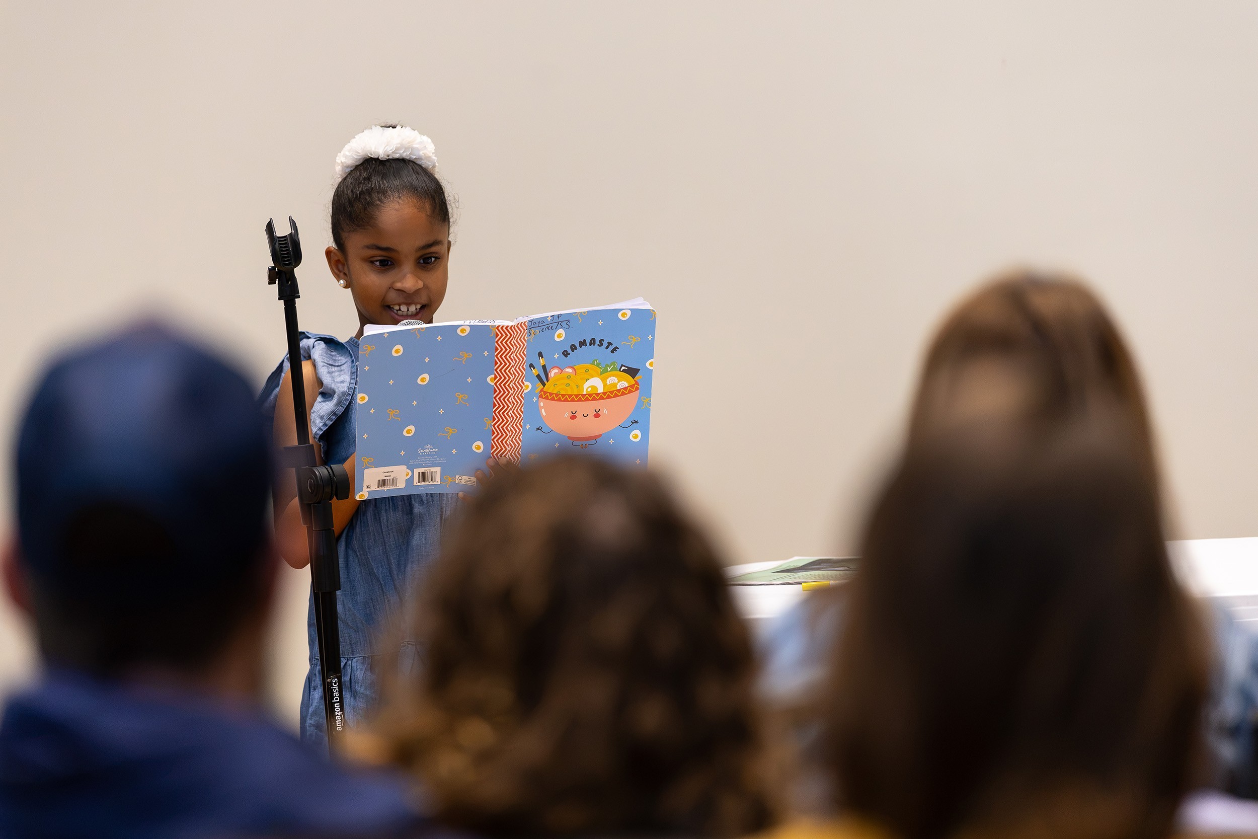 A young girl with a white bow in her hair reads from a book at a microphone in front of an audience.