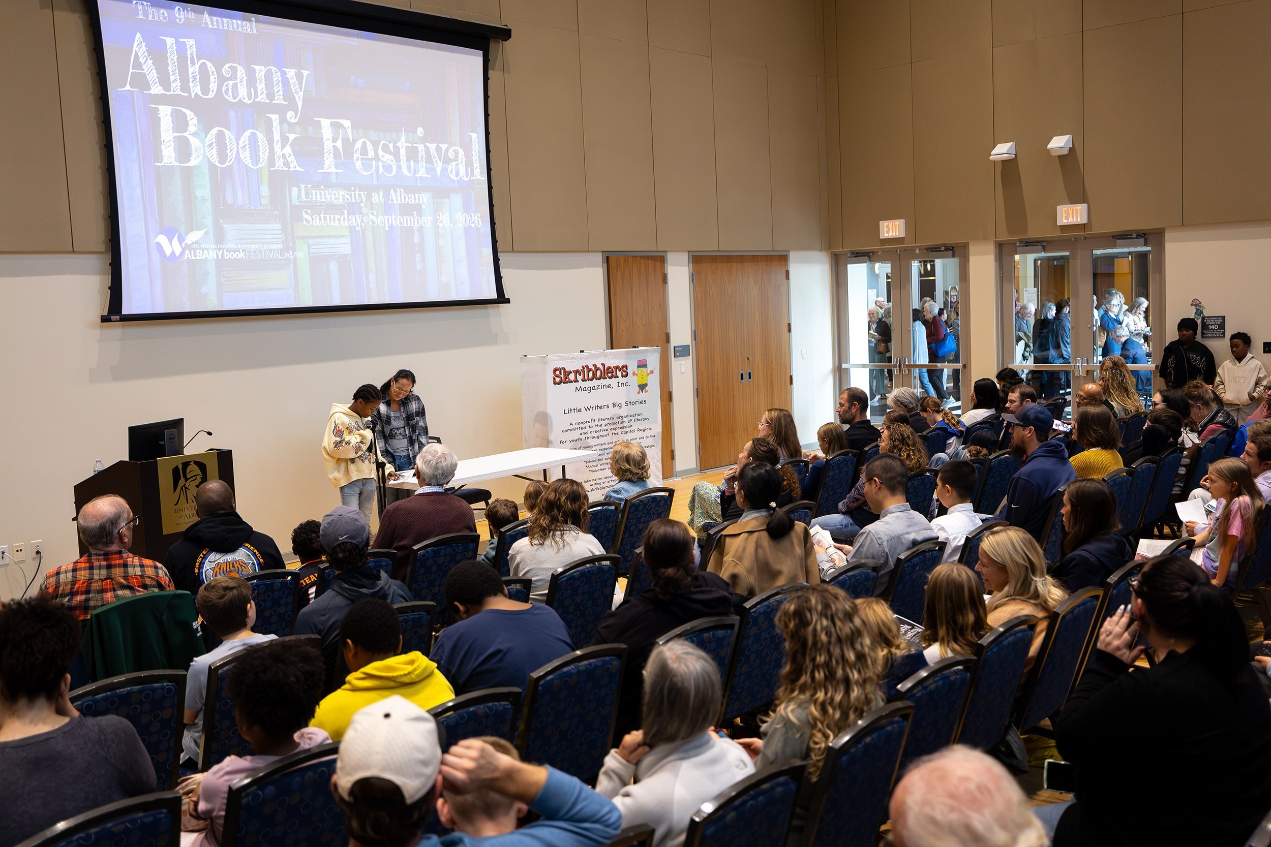 A woman stands near a stage holding a microphone in front of a young girl as she reads a book in front of an audience.