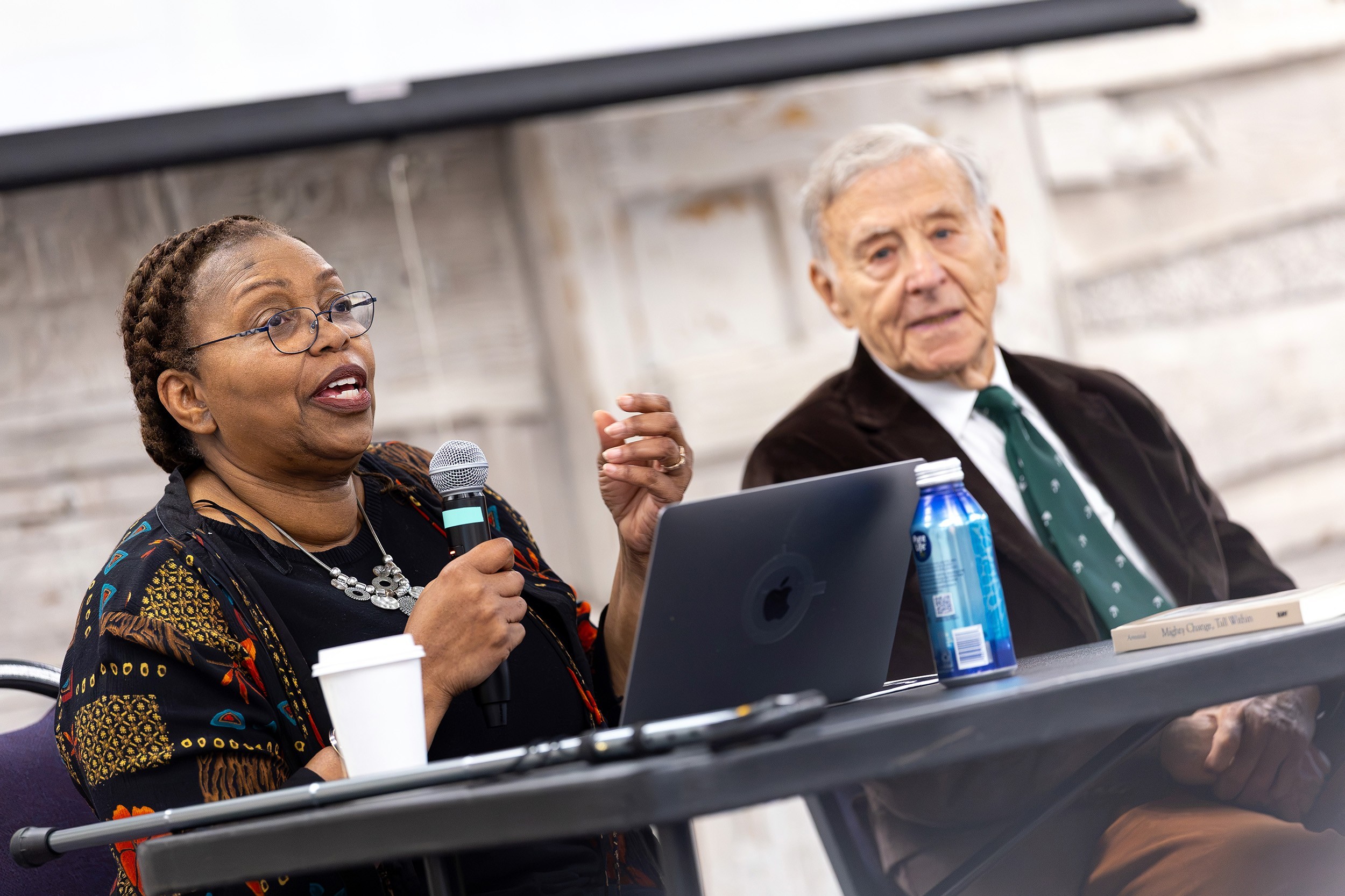 An African American woman and an older white man in a suit sit a table in front of an audience.
