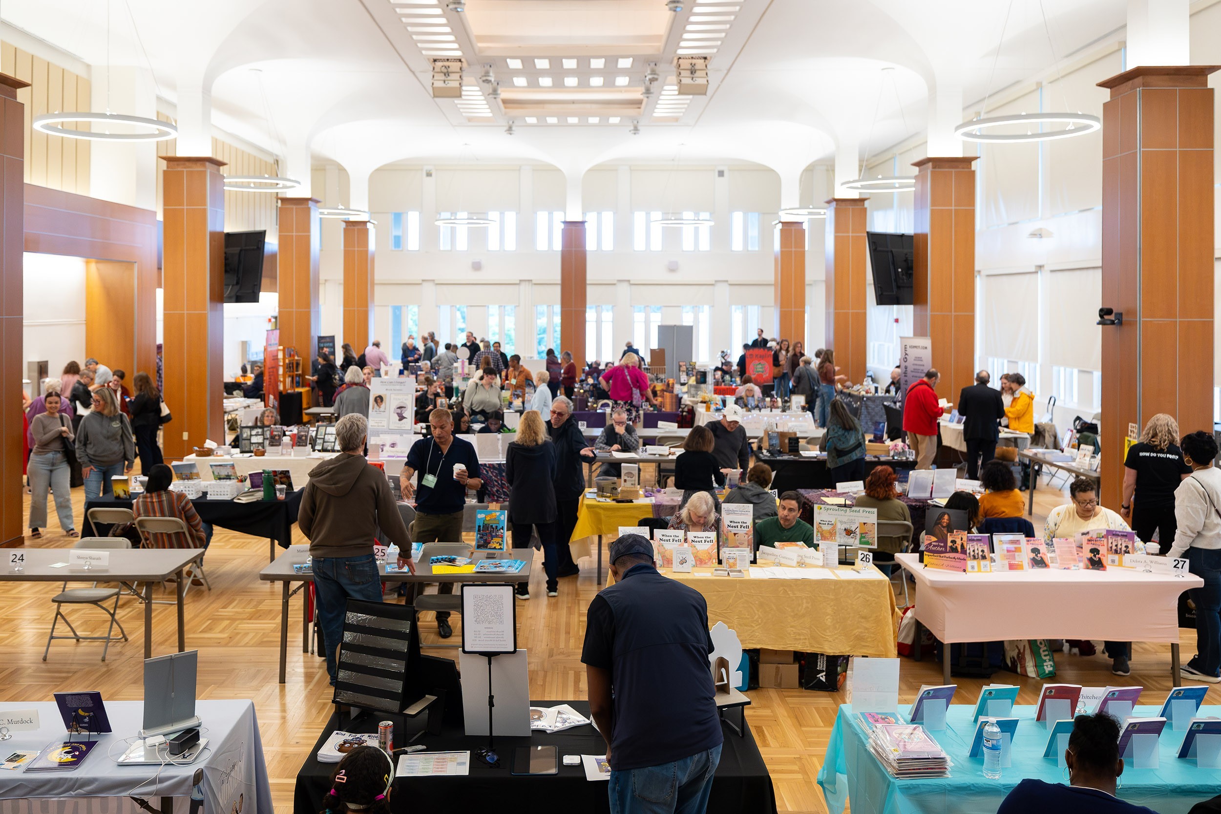 A large group of people move between aisles of people seated with their books during a book show in an auditorium