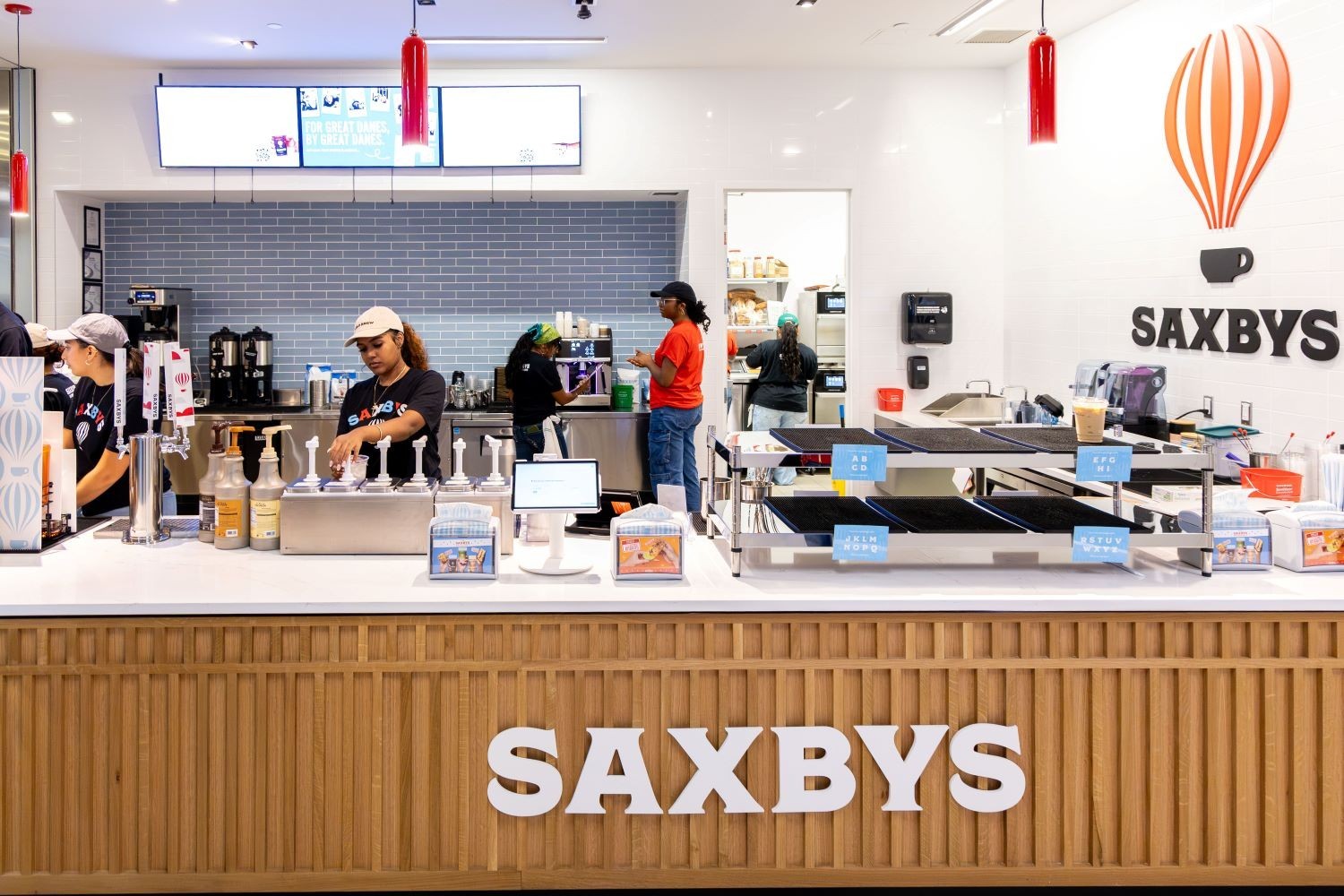 Three students work behind the counter of a coffee eatery with the name SAXBYS in capital letters underneath the counter