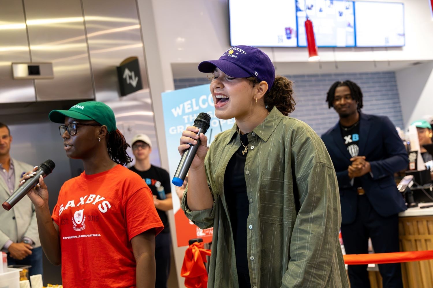 Two young women, one in a red shirt and another in a gray buttoned shirt and hat, sing in front of a new cafe opening inside the Massry School of Business