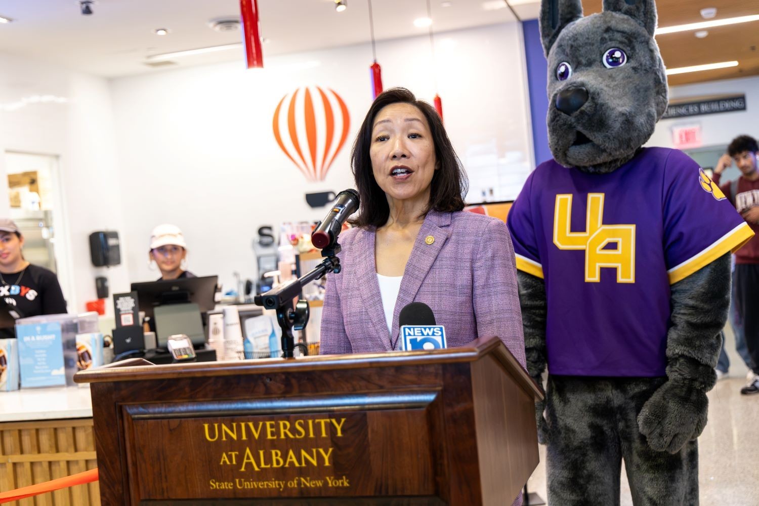 A woman in a purple pant suit with straight dark hair stands at a podium as Damian the Great Dane mascot of UAlbany stands behind her inside the Massry School of Business
