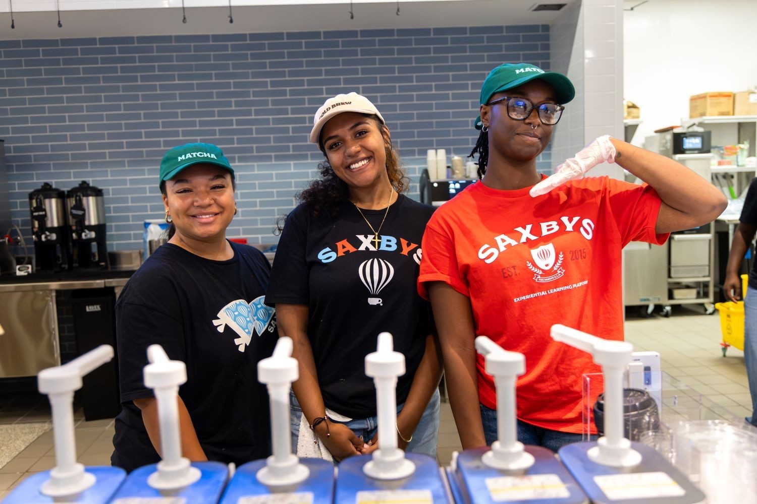 Three students stand behind flavor shot bottles inside a new Saxbys cafe within UAlbany's Massry School of Business
