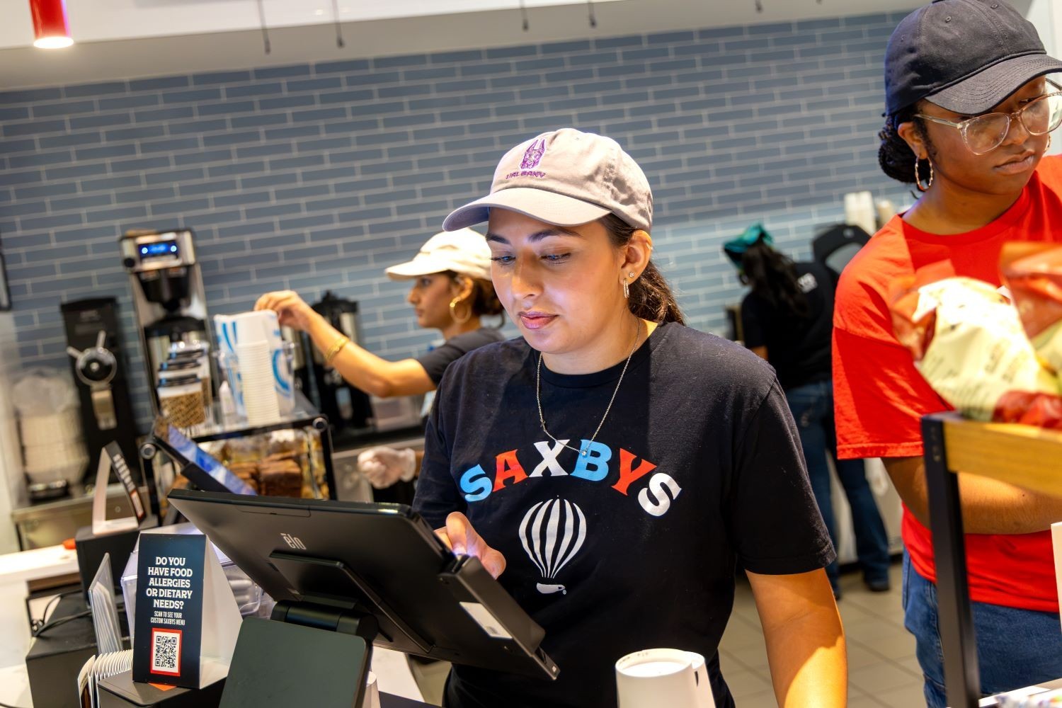 A student wearing a hat and shirt that says Saxbys works at a register insidy the new location at UAlbany's Massry School of Business