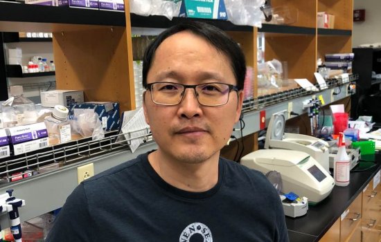 BK Lee stands in a lab with brown shelves behind him containing lab supplies.