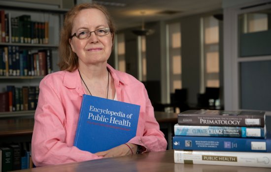 Sue sits at a table in the library. She is wearing a pink blouse and holds an encyclopedia of public health.
