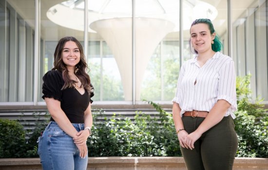 Two young women stand, both with hands clasped in front of them, in front of series of windows