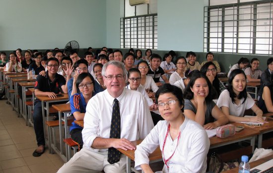 A middle-aged caucasian man sits in classroom with 40 Asian young adults, sitting at school desks