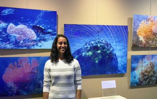 A smiling Sujata Murty, in a gray and white striped sweater, stands in front of several large, underwater photos of a diver near coral formations