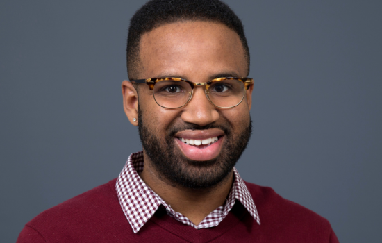 Brandon Lewis smiles at the camera. He is wearing glasses and a red shirt with a checkered collar.