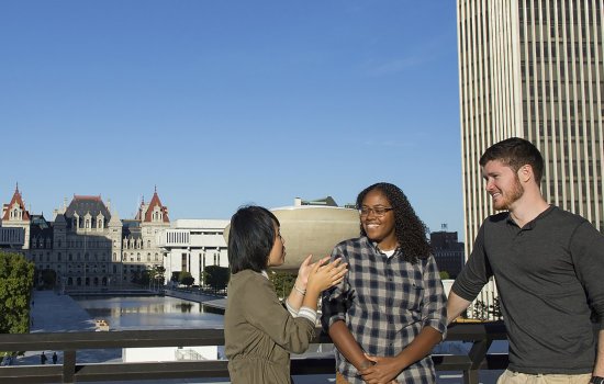 Three students talking outside on a sunny day at the NYS Plaza overlooking the NYS Capital building.