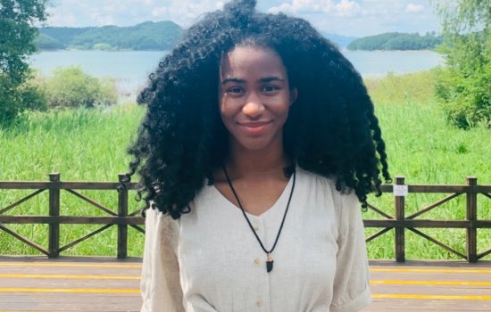 A young woman stands on a large wooden deck with a field, lake and mountains behind her
