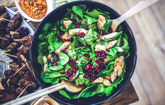 A bowl of salad greens sits on the corner of a table in a black bowl. Pomegranate seeds and chicken sit on top of the leaves.