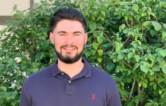CDPHP Scholar, CJ Gil, outside in front of greenery. He has dark hair, facial hair, and is wearing a navy blue polo.