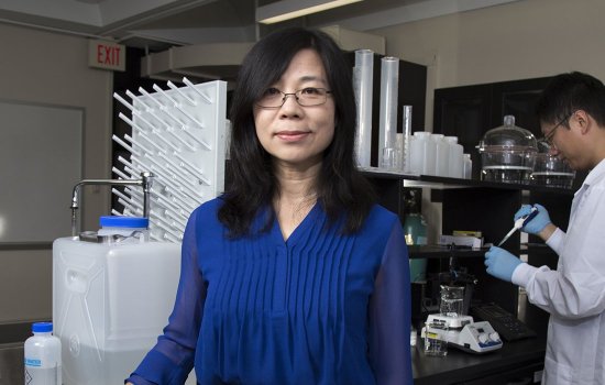 UAlbany Professor of Environmental and Sustainable Engineering Yanna Liang stands in her lab in front of equipement as one of her researchers uses a pipette in the background.