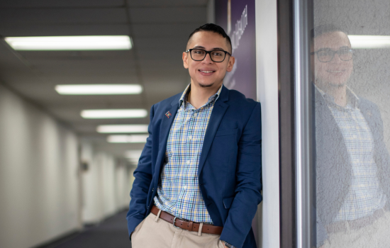 Andrei Chell stands against a wall inside the UAlbany School of Public Health, wearing a blazer and a checkered shirt. He smiles at the camera.
