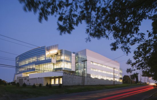 An exterior shot of the Cancer Research Center. The buildign is a pale blue grey with a fully glass wall. Bright lights shine through the glass windows. Next to the center, the road is lit up in a bright red. A tree is in the right corner.