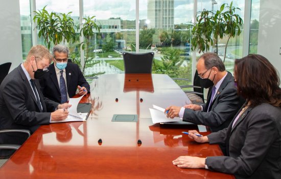 Four people in facemasks and business attire sign papers at a long wooden table in front of a large window with house plants in front of it.