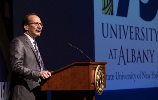 PresidentRodríguez, in a gray suit and purple striped tie, stands at a podium with the words University at Albany projected on a screen to his left. 