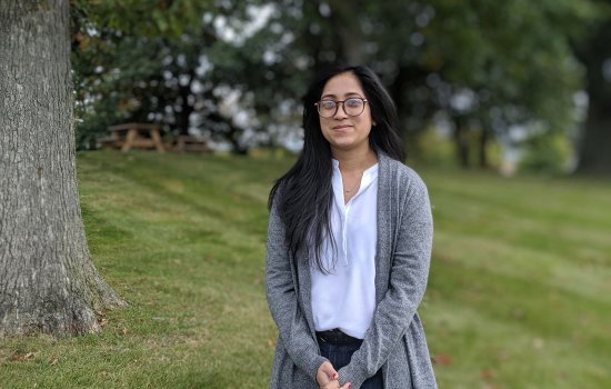 Thoin Begum, a PhD student, stands outside in a field, looking at the camera. Picnic tables and trees are in the background.