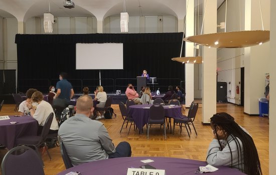 People sit around 5 round tables with purple cloths, listening to a woman speaking from a podium