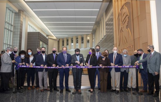 18 people in business clothes stand with scissors, ready to cut a purple ribbon stretched in front of them with the word ETEC repeated on it
