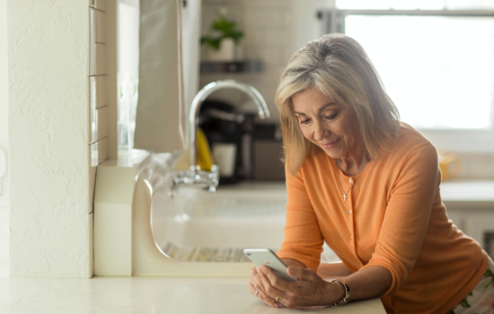 A woman wearing an orange shirt looks at her cell phone in her kitchen. 
