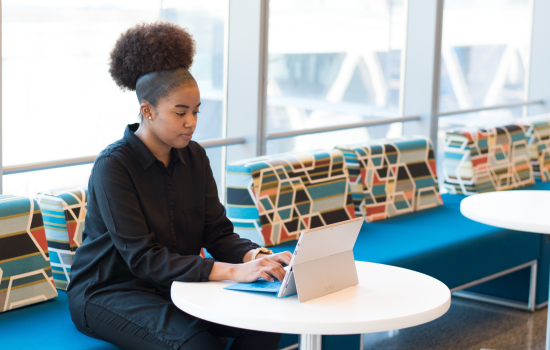 A woman sits at a table, looking at a white tablet propped up next to a keyboard.
