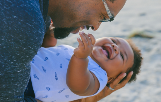 A dad wearing glasses holds his baby on her back, cradling her as she laughs.