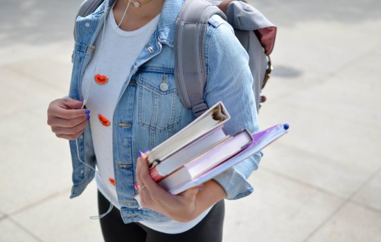 A girl holds three books and a notebook, and is wearing a backpack.