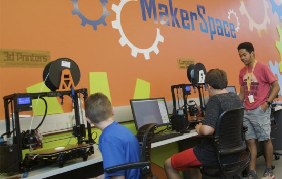 Two children sit at a table with computers and 3D printers are helped by a young man indoors, with a sign MakerSpace on the wall.