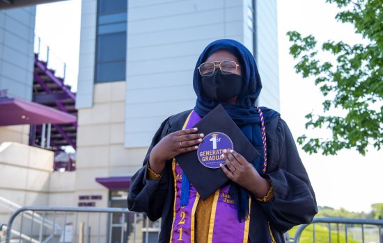 A soman wearing a headscarf, face mask and glasses along with graduation robes holds a circular sticker that says 1st generation graduate.