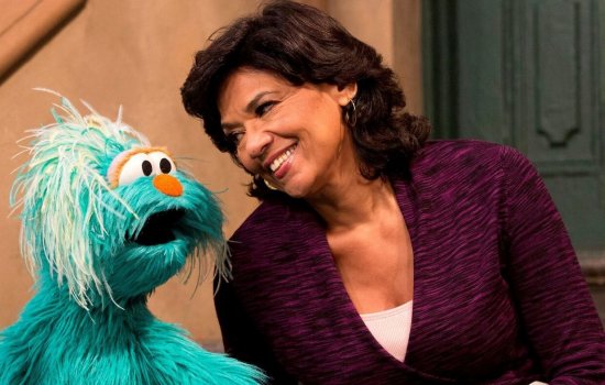 Sonia Manzano, in a purple sweater, leans in and smiles at a large, fuzzy, light blue Muppet