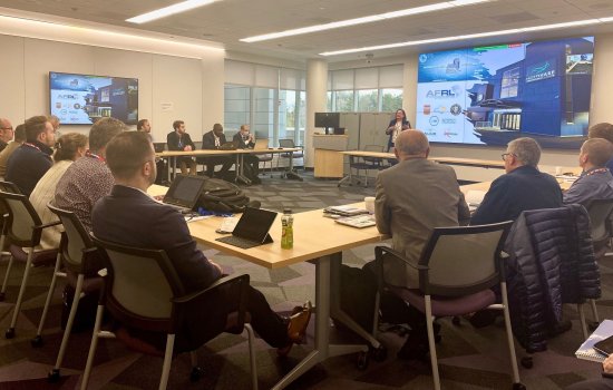 Attendees sit at tables at the circumference of a conference room as a woman standing near a projection screen speaks from the front of the room