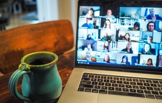A laptop is open on a table and shows a Zoom screen with many participants. Next to the laptop is a mug.