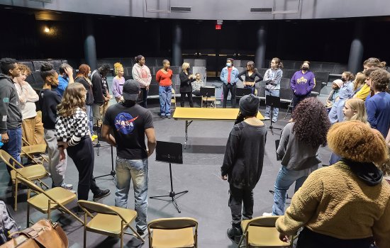 In a theatre in the round, about 30 students in street garb gather in a semi-circle around an older woman giving directions