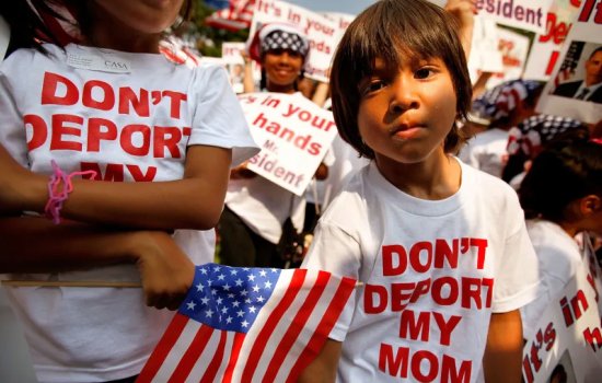 A young boy stands in front of a crowd of children, all wearing Don't Deport My Mom teeshirts.